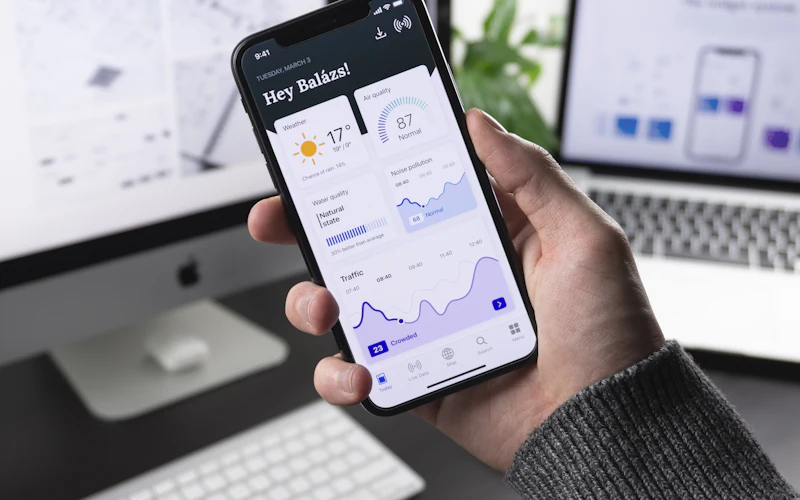 Hand holding a smartphone with a dashboard app in front of a computer